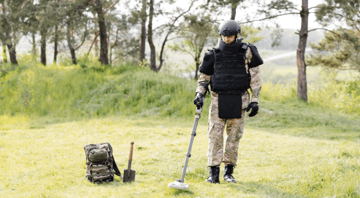 Soldier in protective gear using a metal detector for landmine detection in a grassy field.