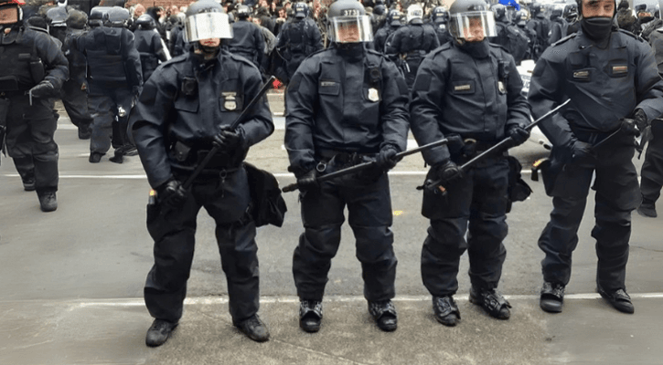 Riot police officers wearing helmets, visors, and tactical armor standing in formation with batons during a public protest or crowd control operation.