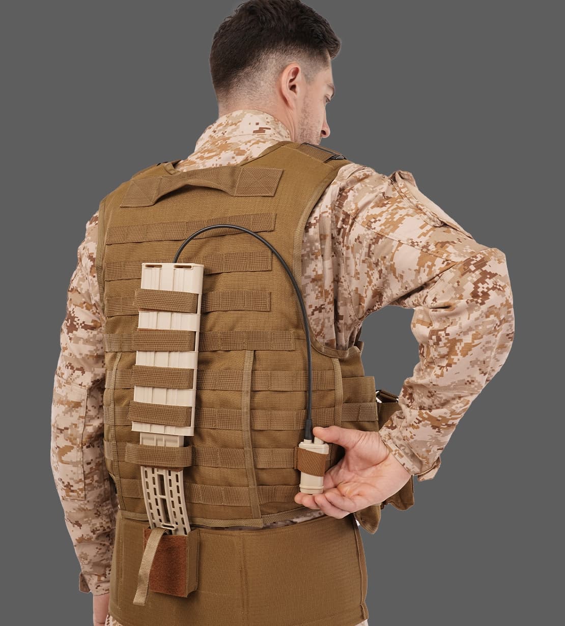 Soldier wearing a tactical vest with MOLLE attachment, holding a connected device on the back panel.